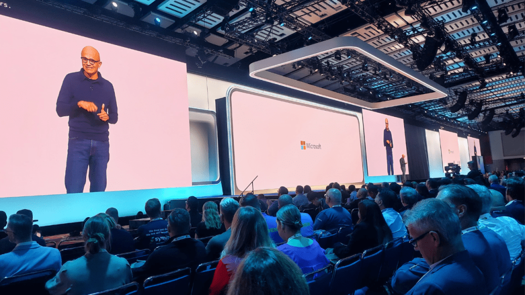 Palco do com 3 telões e publico iluminado pela luz azul assistindo a palestra do CEO da Microsoft