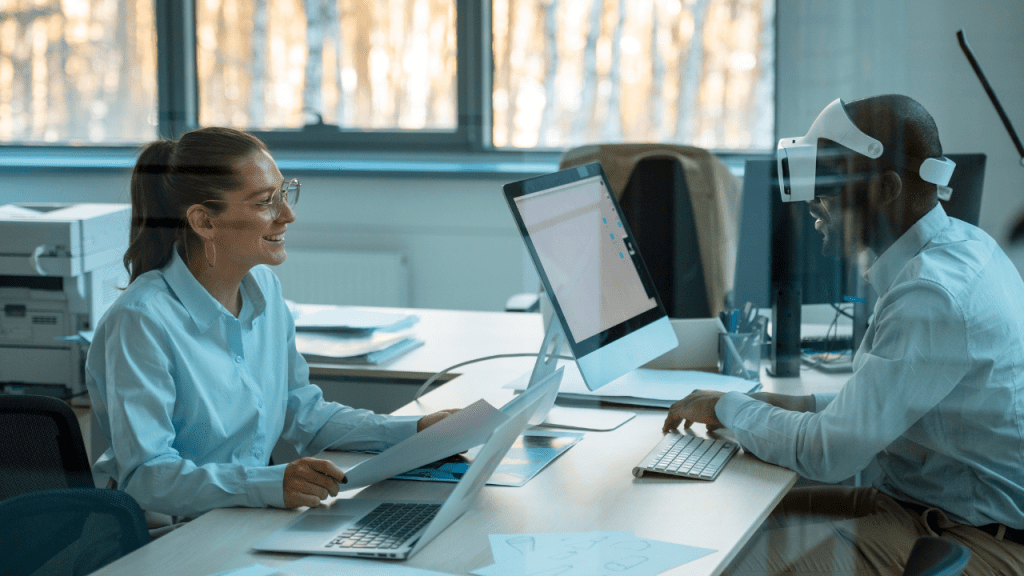 Dois profissionais colaboram em um ambiente de escritório moderno. À esquerda, uma mulher sorridente segura documentos e trabalha em um computador. À direita, um homem utiliza um visor de realidade virtual enquanto interage com outro computador. A cena reflete inovação e tecnologia no ambiente de trabalho.