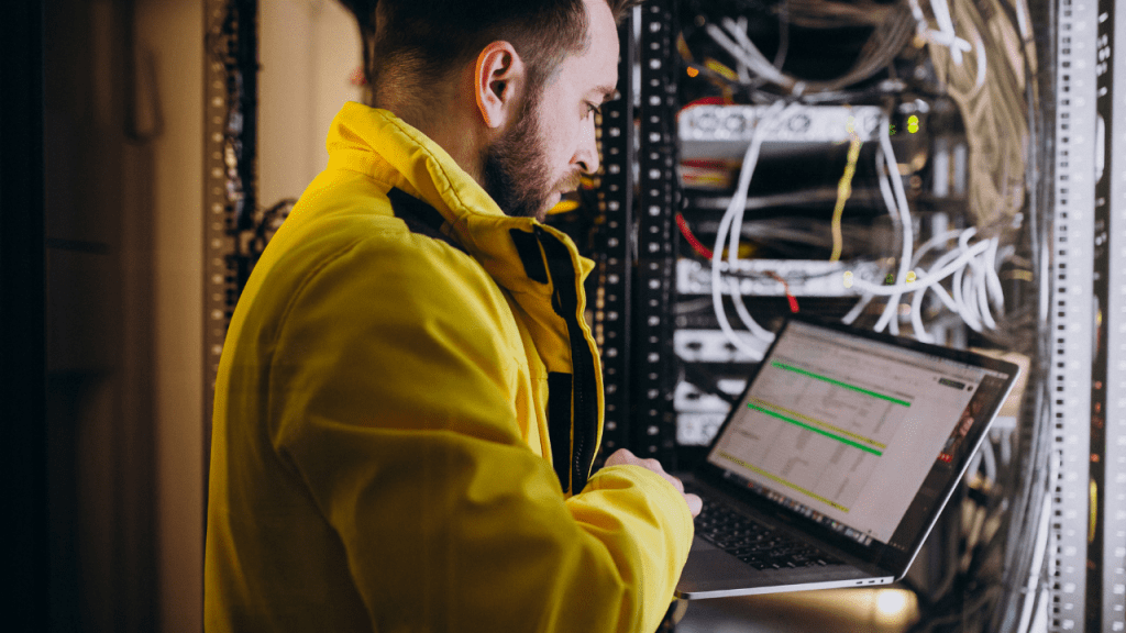 Profissional de TI utilizando um laptop em um data center, cercado por racks de servidores e cabos, vestindo um casaco amarelo.