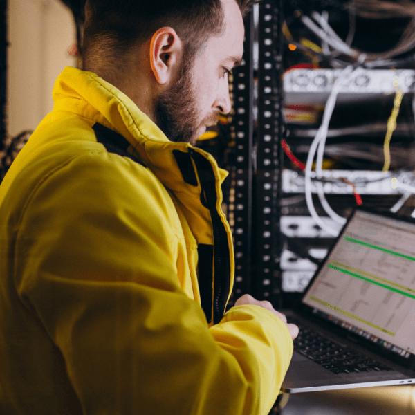 Profissional de TI utilizando um laptop em um data center, cercado por racks de servidores e cabos, vestindo um casaco amarelo.